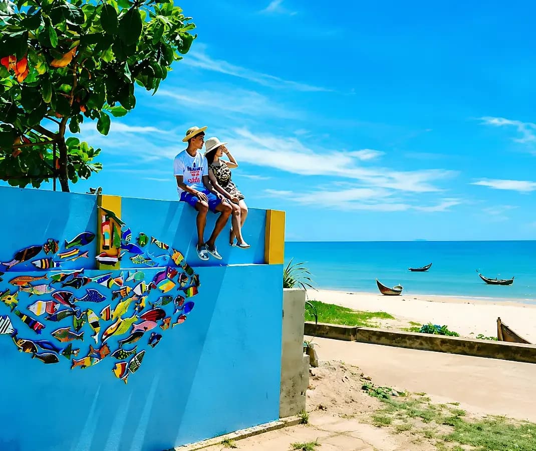 Two young travelers sitting on a wall painted with fish, looking out over Tam Thanh beach